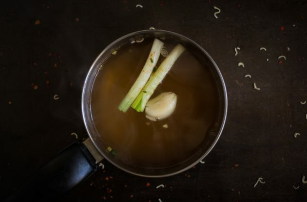 Kā pagatavot mājās gatavotu ramenu: uzvāra buljonu un ievieto tajā ķiploka daiviņu, nomizotu ingveru un zaļo sīpolu kātiņus